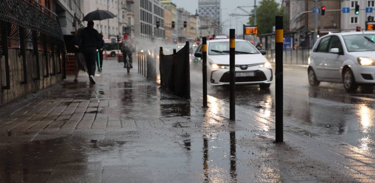 Mokra od deszczu główna przelotowa ulica Trójmiasta (ul Grunwaldzka w Gdańsku) w pochmurny dzień. Zdjęcie z żabiej perspektywy. Na pierwszym planie znajduje się wilgotny, lśniący chodnik, w którym odbijają się światła samochodów i stojące wzdłuż krawężnika czarne słupki z żółtymi paskami. Po prawej stronie, po zalanej jezdni, jadą samochody (w tym biała taksówka), rozbryzgując wodę. Po lewej stronie chodnikiem idą piesi, z których jeden niesie rozłożony czarny parasol