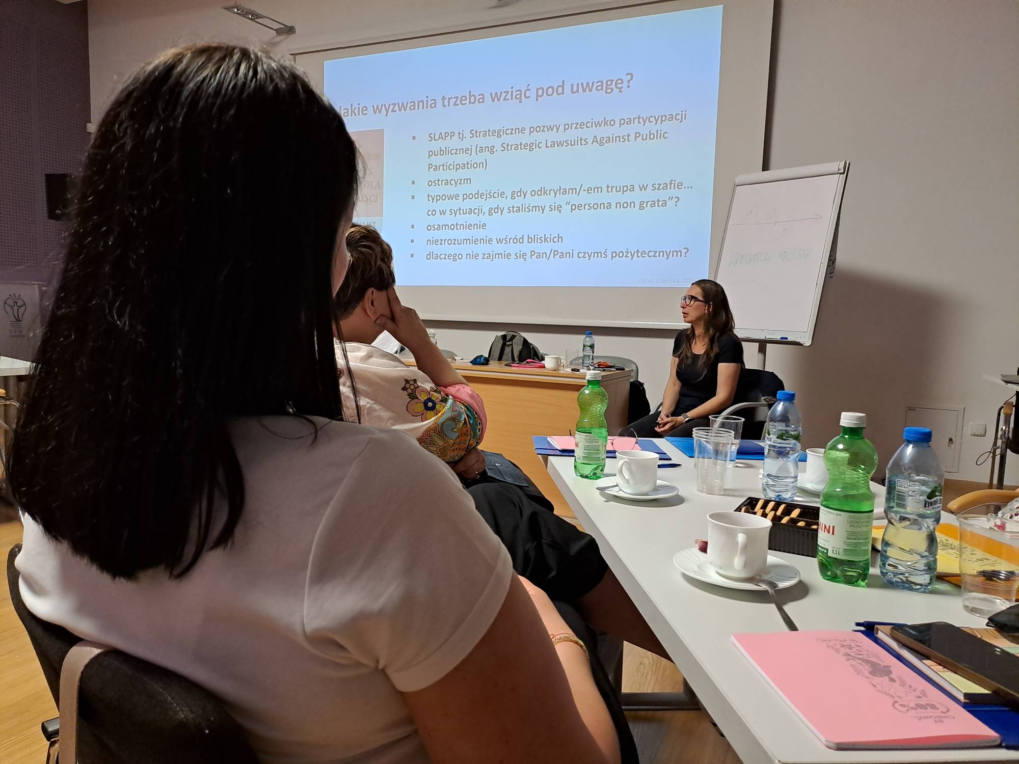 Zdjęcie wykonane z perspektywy zza pleców uczestniczki warsztatów dla liderek w Chojnicach, patrzącej w stronę ekranu projekcyjnego. Na ekranie wyświetlony jest slajd o tytule „Jakie wyzwania trzeba wziąć pod uwagę?” z wypunktowanymi zagadnieniami takimi jak SLAPP, ostracyzm, osamotnienie czy niezrozumienie wśród bliskich. Po prawej stronie ekranu, obok tablicy typu flipchart, siedzi prelegentka Iwona Janicka w okularach i ciemnym t-shircie. W dolnej części kadru widoczny jest stół z filiżankami kawy, butelkami wody mineralnej oraz różowym notesem