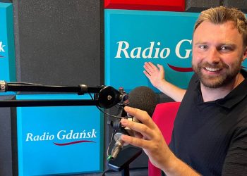 Uśmiechnięty mężczyzna z krótkim zarostem i w ciemnej koszulce siedzi w studiu radiowym przy mikrofonie, wskazując dłonią na turkusowy panel z logo „Radio Gdańsk” w tle. To Wojciech Czajka, fizjoterapeuta i osteopata, szef słupskiego Centrum Rehabilitacji Avenir. Na antenie mówił o tym, jak przygotować się do wakacyjnych wypraw i długich wędrówek