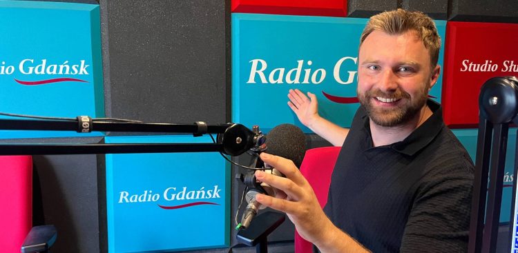 Uśmiechnięty mężczyzna z krótkim zarostem i w ciemnej koszulce siedzi w studiu radiowym przy mikrofonie, wskazując dłonią na turkusowy panel z logo „Radio Gdańsk” w tle. To Wojciech Czajka, fizjoterapeuta i osteopata, szef słupskiego Centrum Rehabilitacji Avenir. Na antenie mówił o tym, jak przygotować się do wakacyjnych wypraw i długich wędrówek