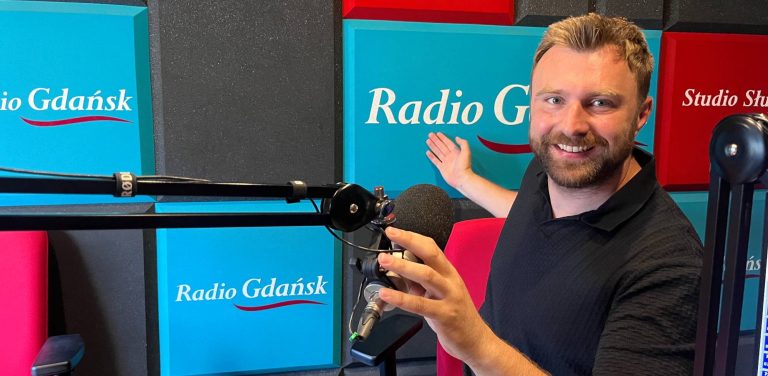 Uśmiechnięty mężczyzna z krótkim zarostem i w ciemnej koszulce siedzi w studiu radiowym przy mikrofonie, wskazując dłonią na turkusowy panel z logo „Radio Gdańsk” w tle. To Wojciech Czajka, fizjoterapeuta i osteopata, szef słupskiego Centrum Rehabilitacji Avenir. Na antenie mówił o tym, jak przygotować się do wakacyjnych wypraw i długich wędrówek