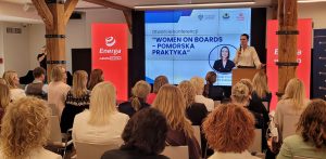 Widok od tyłu na salę konferencyjną wypełnioną publicznością, w której przeważają kobiety. Na scenie po prawej stronie, za drewnianą mównicą, stoi kobieta w jasnej bluzce (wojewoda pomorska Beata Rutkiewicz) i przemawia do mikrofonu. W tle znajduje się duży ekran wyświetlający slajd z napisem „Otwarcie konferencji 'WOMEN ON BOARDS – POMORSKA PRAKTYKA'” oraz portretem wojewody. Po obu stronach ekranu stoją wysokie, czerwone banery reklamowe z białym logo i napisem „Energa GRUPA ORLEN”