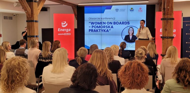 Widok od tyłu na salę konferencyjną wypełnioną publicznością, w której przeważają kobiety. Na scenie po prawej stronie, za drewnianą mównicą, stoi kobieta w jasnej bluzce (wojewoda pomorska Beata Rutkiewicz) i przemawia do mikrofonu. W tle znajduje się duży ekran wyświetlający slajd z napisem „Otwarcie konferencji 'WOMEN ON BOARDS – POMORSKA PRAKTYKA'” oraz portretem wojewody. Po obu stronach ekranu stoją wysokie, czerwone banery reklamowe z białym logo i napisem „Energa GRUPA ORLEN”