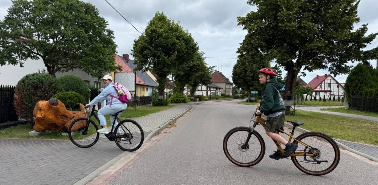 Kobieta z różowym plecakiem i chłopiec w czerwonym kasku jadą na rowerach po asfaltowej drodze w miejscowości Smołdzino. W tle widać tradycyjne domy, drzewa, drogowskaz do Kluk oraz drewnianą rzeźbę leśnego zwierzęcia na trawniku. Przywrócenie ruchu rowerowego na ścieżce Słowińskiego Parku Narodowego prowadzącej do Wydmy Łąckiej