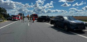 Miejsce kolizji drogowej na wielopasmowej autostradzie A1. Na asfalcie rozsypane są liczne odłamki i elementy uszkodzonych karoserii. Po prawej stronie stoją dwa uszkodzone, ciemne samochody typu SUV – jeden z nich stoi ukośnie w poprzek pasa ruchu. Po lewej stronie jezdni zaparkowane są czerwone wozy straży pożarnej oraz inne pojazdy ratownicze, przy których pracują strażacy oraz ratownik medyczny