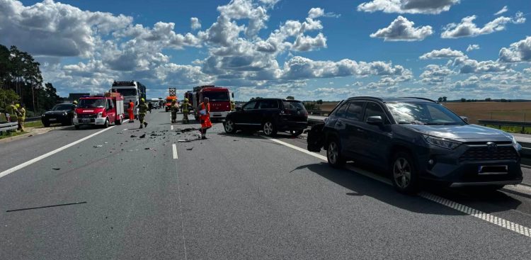 Miejsce kolizji drogowej na wielopasmowej autostradzie A1. Na asfalcie rozsypane są liczne odłamki i elementy uszkodzonych karoserii. Po prawej stronie stoją dwa uszkodzone, ciemne samochody typu SUV – jeden z nich stoi ukośnie w poprzek pasa ruchu. Po lewej stronie jezdni zaparkowane są czerwone wozy straży pożarnej oraz inne pojazdy ratownicze, przy których pracują strażacy oraz ratownik medyczny