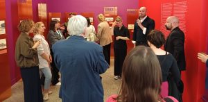 Wystawa „Opera! 100 lat projektowania Opery w Gdańsku” w Gdańskim Centrum Sztuki Współczesnej. Grupa osób słuchających przemawiającego mężczyzny z mikrofonem w nowoczesnej sali wystawowej o intensywnie fioletowych i czerwonych ścianach. W tle widoczne są pomarańczowe, pionowe tablice ekspozycyjne z datami „1939”, „1911” oraz „1899” i rysunkami architektonicznymi.