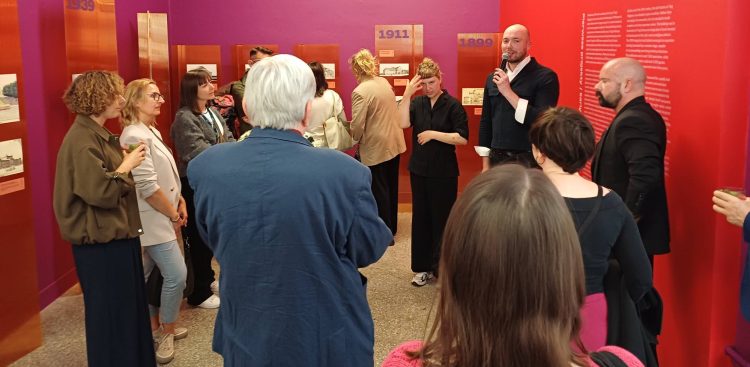 Wystawa „Opera! 100 lat projektowania Opery w Gdańsku” w Gdańskim Centrum Sztuki Współczesnej. Grupa osób słuchających przemawiającego mężczyzny z mikrofonem w nowoczesnej sali wystawowej o intensywnie fioletowych i czerwonych ścianach. W tle widoczne są pomarańczowe, pionowe tablice ekspozycyjne z datami „1939”, „1911” oraz „1899” i rysunkami architektonicznymi.