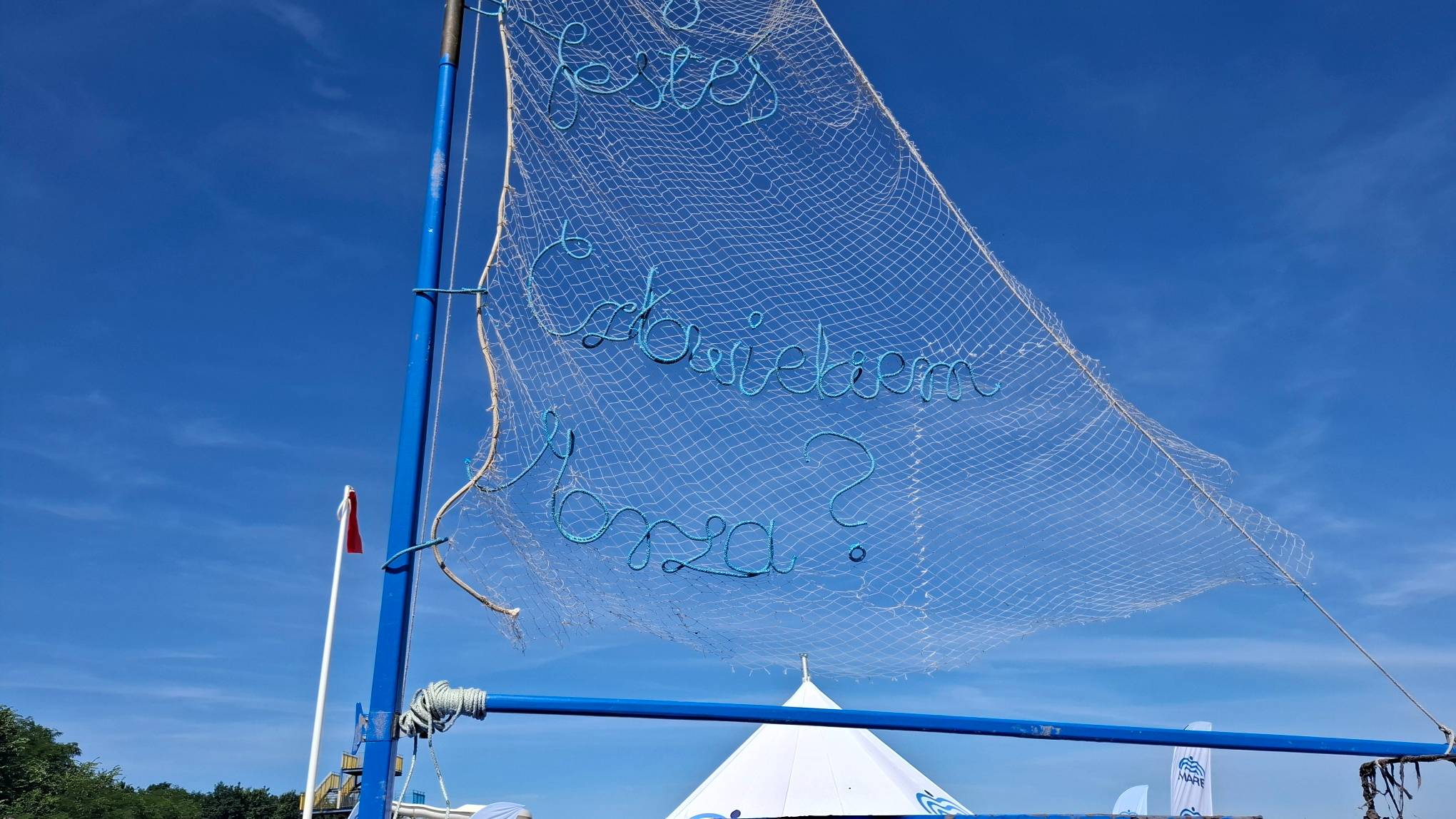 Instalacja artystyczna na plaży w Gdańsku-Brzeźnie. Zbliżenie na transparentny żagiel wykonany z białej sieci rybackiej, rozpięty na niebieskim maszcie na tle czystego, niebieskiego nieba. Z niebieskiego sznura wplecionego w sieć ułożono napis w formie pytania: "Czy jesteś Człowiekiem Morza?". W dolnej części kadru widać szczyt białego namiotu