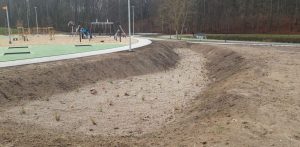 Szerokie, ziemno-piaszczyste zagłębienie (mulda chłonna) z rzadkimi, młodymi kępkami roślin na dnie. Wzdłuż krawędzi zagłębienia biegnie jasny, zakrzywiony chodnik, za którym znajduje się nowoczesny plac zabaw z zieloną nawierzchnią poliuretanową i urządzeniami rekreacyjnymi. W tle widoczne są drzewa i ściana lasu. Gdynia: Infrastruktura wybudowana w ramach I i II etapu zadania inwestycyjnego nr 8 „Zagospodarowanie terenów przy ul. Wielkokackiej i ul. 2 Morskiego Pułku Strzelców w Gdyni” z zastosowaniem rozwiązań opartych na przyrodzie, objętego projektem „Rozwój systemu gospodarowania wodami opadowymi na terenie Gdyni - część IV