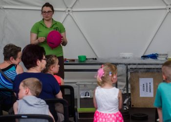 Kobieta w zielonej koszulce i okularach prowadzi pokaz naukowy w sferycznym namiocie, trzymając w dłoniach różowy balon. Przed nią na krzesłach siedzi grupa dzieci i dorosłych, z których część zgłasza się, unosząc ręce. Po prawej stronie znajduje się stół z akcesoriami do doświadczeń, z którego unosi się gęsta, biała para, a obok stoi skrzynia z napisem „W krainie ciekłego mrozu”. To zlot gdańskich rodzin zastępczych w Centrum Hevelianum w Gdańsku