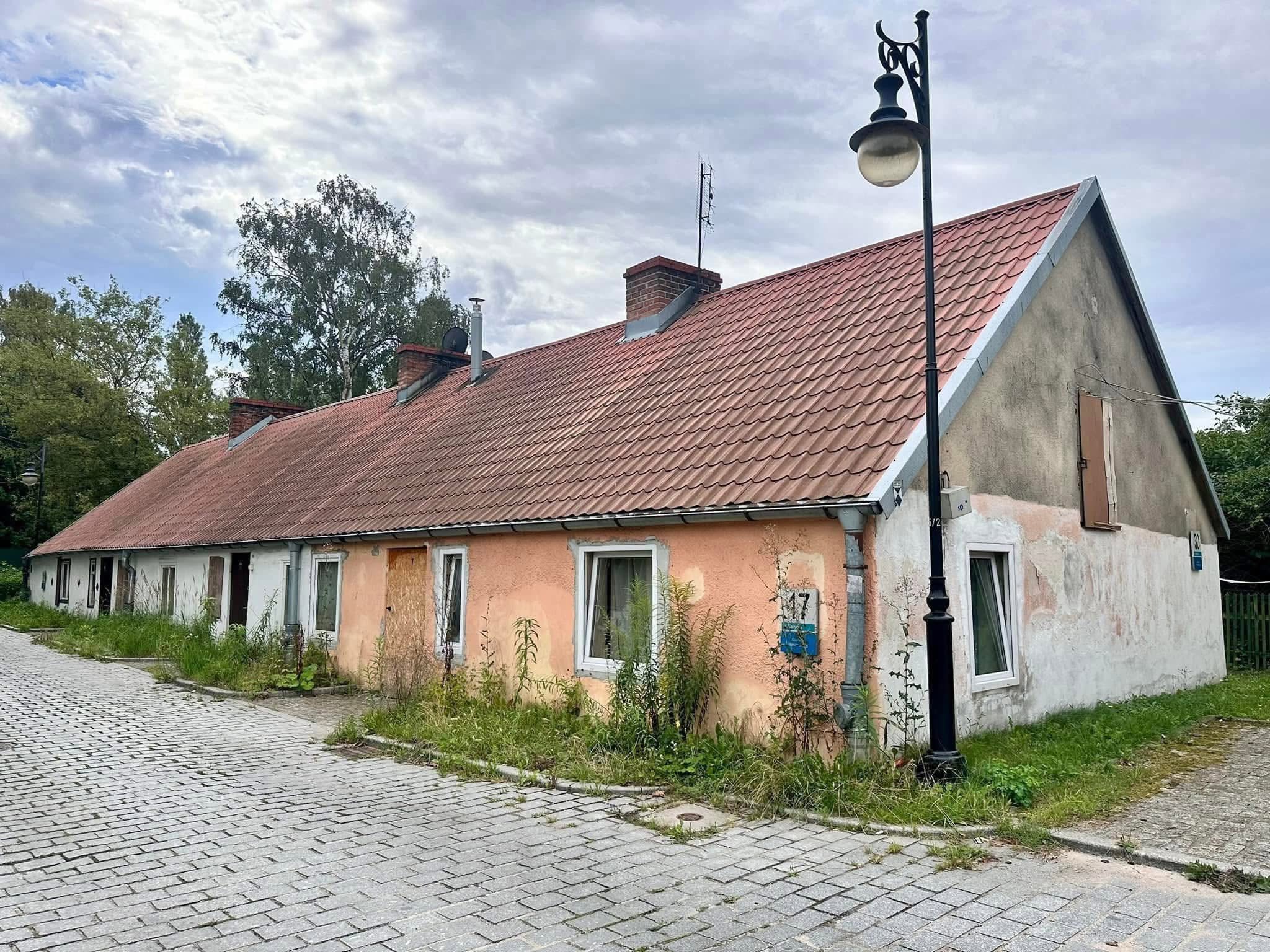 Parterowa chata rybacka (checz rybacka) o spadzistym dachu z czerwoną dachówką i łososiowo-różową oraz białą elewacją przed zbliżającym się remontem. Przed budynkiem, przy brukowanej ulicy, stoi czarna, retro latarnia uliczna. Na ścianie przy oknie widnieje tabliczka adresowa z numerem 17. U podstawy budynku rosną wysokie chwasty