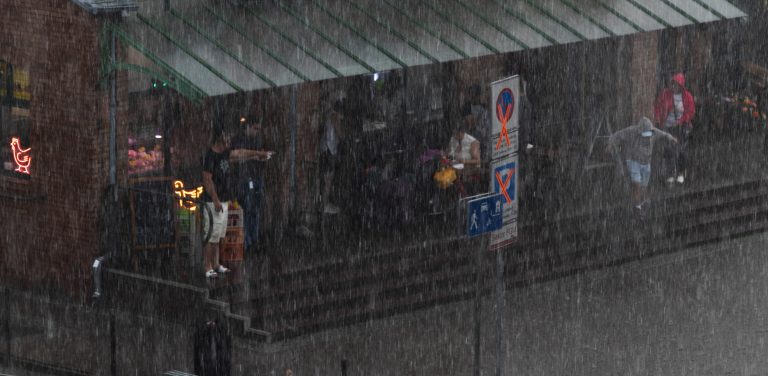 Ludzie chroniący się pod szklanym zadaszeniem budynku z ciemnej cegły podczas gwałtownej ulewy w Gdańsku. Na pierwszym planie widoczne są gęste, pionowe strugi intensywnego deszczu. Miasto informuje, że na inwestycje przeciwpowodziowe wydało 675 mln zł w 5 lat