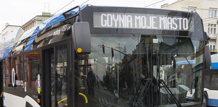 Niebiesko-biały autobus komunikacji miejskiej w Gdyni (ZKM Gdynia) jedzie miejską ulicą. W tle widoczne są drzewa, chodnik oraz fragmenty budynków mieszkalnych
