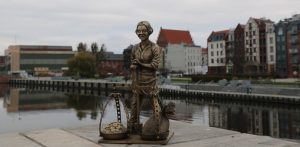 Mosiężna figurka elbląskiego Piekarczyka z wagą szalkową, ustawiona na betonowym nabrzeżu na tle rzeki Elbląg i staromiejskich kamienic. W Elblągu odbędą się Dni Morza, które mają przypominać o morskiej tradycji miasta
