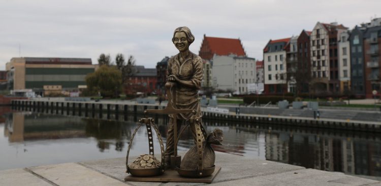 Mosiężna figurka elbląskiego Piekarczyka z wagą szalkową, ustawiona na betonowym nabrzeżu na tle rzeki Elbląg i staromiejskich kamienic. W Elblągu odbędą się Dni Morza, które mają przypominać o morskiej tradycji miasta