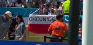 Kadr z transmisji telewizyjnej przedstawiający polską biało-czerwoną flagę z dużym czarnym napisem „CHOJNICE” umieszczonym na białym tle. Flaga jest zawieszona na barierki odgradzającej trybuny stadionu. Na pierwszym planie widoczni są pracownicy obsługi i ochrony: po lewej stronie dwie osoby w szarych kamizelkach, a przed samą flagą tyłem stoi steward w pomarańczowej koszulce. W tle widać zarys innych osób na trybunach