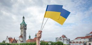 Fotografia przedstawia dłoń trzymającą dwie małe flagi Ukrainy na tle charakterystycznego budynku sopockiego domu zdrojowego z wieżą. Obraz jest ilustracją do informacji o podpisaniu przez władze Sopotu rezolucji przeciwko nastrojom antyukraińskim