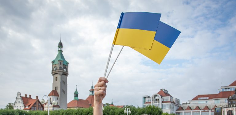 Fotografia przedstawia dłoń trzymającą dwie małe flagi Ukrainy na tle charakterystycznego budynku sopockiego domu zdrojowego z wieżą. Obraz jest ilustracją do informacji o podpisaniu przez władze Sopotu rezolucji przeciwko nastrojom antyukraińskim