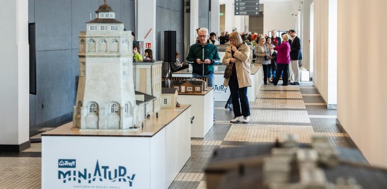Jasny hol wystawy „Trefl Miniatury” w Muzeum Emigracji w Gdyni. Na pierwszym planie po lewej stronie stoi szczegółowa makieta nieistniejącej już Portowej Wieży Ciśnień w skali 1:25, umieszczona na postumencie z logo wystawy. W głębi korytarza odwiedzający oglądają kolejne miniatury przedwojennych budynków