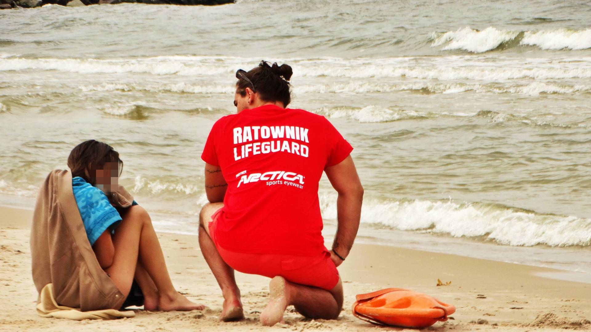 Ratownik w czerwonej koszulce z napisem „Ratownik Lifeguard” klęczy na piaszczystej plaży obok młodej dziewczyny owiniętej kocem i ręcznikiem. Na piasku leży pomarańczowa boja, a w tle widać morskie fale