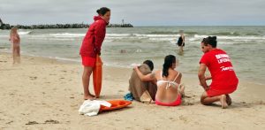 Dwójka ratowników w czerwonych strojach udziela pomocy dwóm osobom siedzącym na piaszczystej plaży nad morzem w Ustce. Jedna z poszkodowanych osób jest owinięta brązowym kocem, obok leży pomarańczowy sprzęt ratunkowy, a w tle widać wzburzone fale