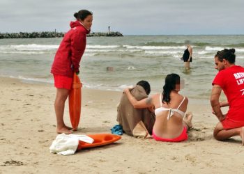 Dwójka ratowników w czerwonych strojach udziela pomocy dwóm osobom siedzącym na piaszczystej plaży nad morzem w Ustce. Jedna z poszkodowanych osób jest owinięta brązowym kocem, obok leży pomarańczowy sprzęt ratunkowy, a w tle widać wzburzone fale