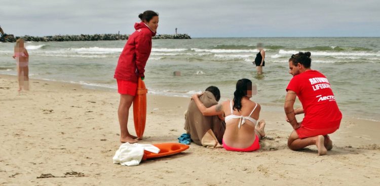 Dwójka ratowników w czerwonych strojach udziela pomocy dwóm osobom siedzącym na piaszczystej plaży nad morzem w Ustce. Jedna z poszkodowanych osób jest owinięta brązowym kocem, obok leży pomarańczowy sprzęt ratunkowy, a w tle widać wzburzone fale