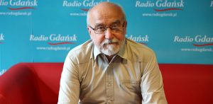 Kapitan Tadeusz Hatalski w Radiu Gdańsk. Mężczyzna siedzi na czerwonej sofie na tle ścianki, na której widać logo rozgłośni "Radio Gdańsk" powtórzone wielokrotnie.