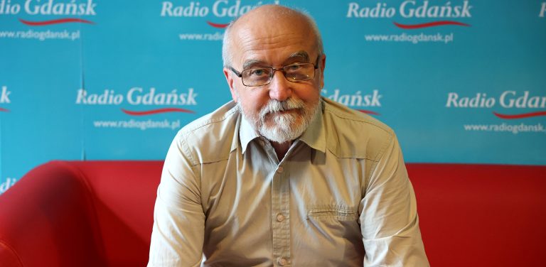 Kapitan Tadeusz Hatalski w Radiu Gdańsk. Mężczyzna siedzi na czerwonej sofie na tle ścianki, na której widać logo rozgłośni "Radio Gdańsk" powtórzone wielokrotnie.
