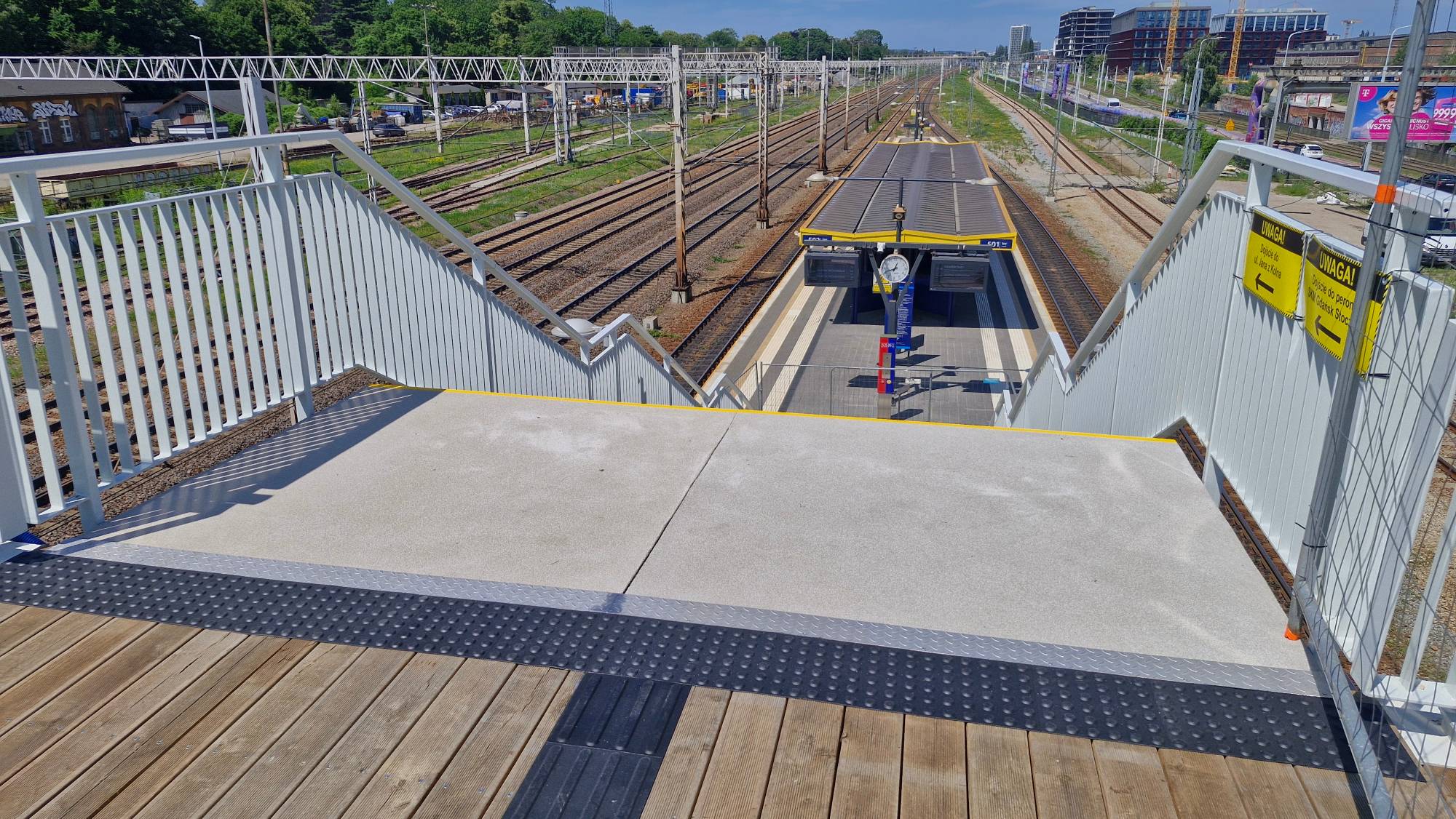 Widok z nowej kładki przy przystanku SKM Gdańsk Stocznia w dół na betonowe schody prowadzące na peron stacji SKM Gdańsk Stocznia. Na pierwszym planie znajduje się drewniany pomost z czarnym pasem ostrzegawczym, a dalej schody z białymi barierkami, na których wiszą dwie żółte tablice informacyjne. W tle widać tory kolejowe, zadaszenie peronu oraz sieć trakcyjną
