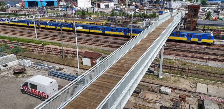 Nowa, stalowa kładka o białej konstrukcji i drewnianej nawierzchni, przebiegająca nad torami kolejowymi. Na torach widoczne są żółto-niebieskie pociągi SKM, a na dole, po lewej stronie, zaparkowany jest biały furgon z czerwonym napisem Strabag. W tle rozciągają się industrialne tereny stoczniowe Gdańska