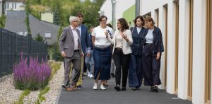 Grupa gdańskich samorządowców, w tym prezydent Aleksandra Dulkiewicz i jej zastępczyni Monika Chabior, idzie chodnikiem wzdłuż nowoczesnego budynku żłobka przy ul. Czerskiej. Osoby na zdjęciu rozmawiają ze sobą i gestykulują. Po prawej stronie widoczna jest biała ściana budynku z dużymi drewnianymi oknami, a po lewej grafitowy płot panelowy, pas jasnego żwiru oraz kępy kwitnących na fioletowo kwiatów. Spotkanie z dziennikarzami dotyczące wydatków na remonty w placówkach edukacyjnych w Gdańsku