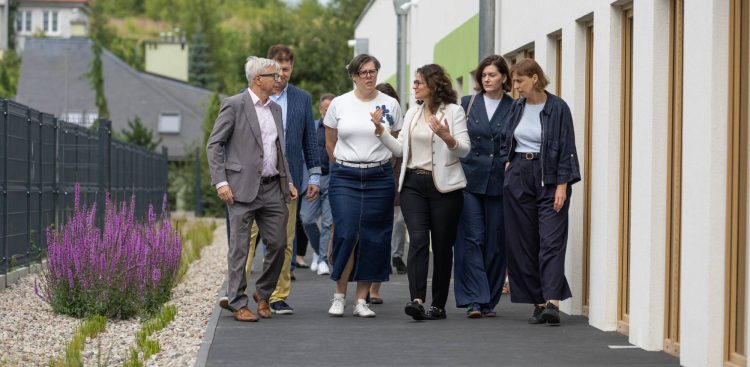 Grupa gdańskich samorządowców, w tym prezydent Aleksandra Dulkiewicz i jej zastępczyni Monika Chabior, idzie chodnikiem wzdłuż nowoczesnego budynku żłobka przy ul. Czerskiej. Osoby na zdjęciu rozmawiają ze sobą i gestykulują. Po prawej stronie widoczna jest biała ściana budynku z dużymi drewnianymi oknami, a po lewej grafitowy płot panelowy, pas jasnego żwiru oraz kępy kwitnących na fioletowo kwiatów. Spotkanie z dziennikarzami dotyczące wydatków na remonty w placówkach edukacyjnych w Gdańsku