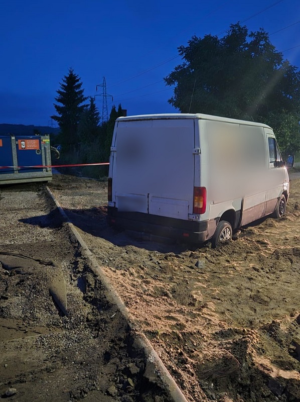 Widok od tyłu na biały samochód dostawczy zakopany w sypkim piasku na placu budowy o zmierzchu w Lęborku. Obok widoczne jest niebieskie urządzenie przemysłowe ogradzające teren taśmą ostrzegawczą. Kierowca utknął, a przed policją schował się "na pace", czyli w części bagażowej pojazdu. 