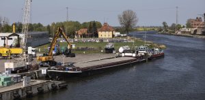 Barka rzeczna zacumowana przy betonowym nabrzeżu portowym oznaczonym numerem 2. Na brzegu stoi żółta koparka przeładunkowa na kołach oraz biały dźwig kratownicowy. W tle widoczne jest koryto rzeki, drzewa, tradycyjny żółty budynek o spadzistym dachu oraz elementy zabudowy przemysłowej po prawej stronie