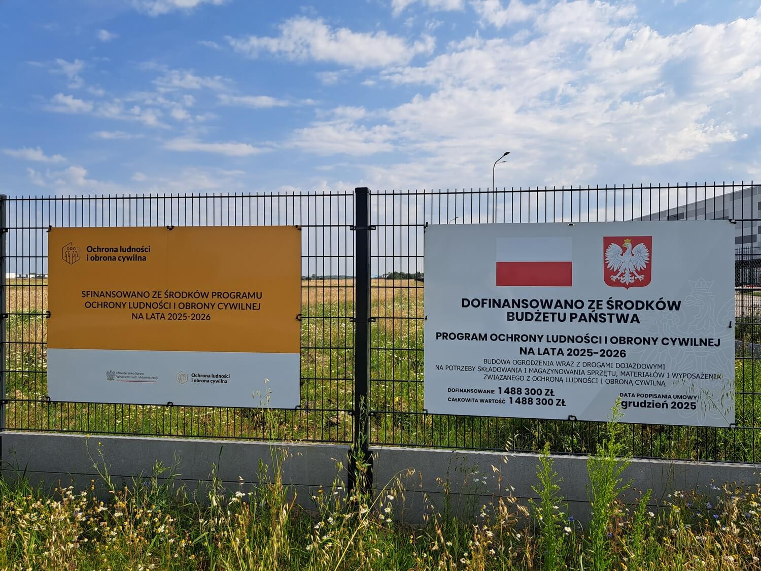 Dwie tablice informacyjne umieszczone na metalowym ogrodzeniu działki. Informują o dofinansowaniu budowy ogrodzenia i dróg dojazdowych z Programu Ochrony Ludności i Obrony Cywilnej na lata 2025–2026 na kwotę blisko 1,5 mln zł. Drogi mogą w przyszłości doprowadzić do mającej powstać nowej remizie, która być może stanie się siedzibą Komendy Wojewódzkiej Państwowej Straży Pożarnej w Gdańsku