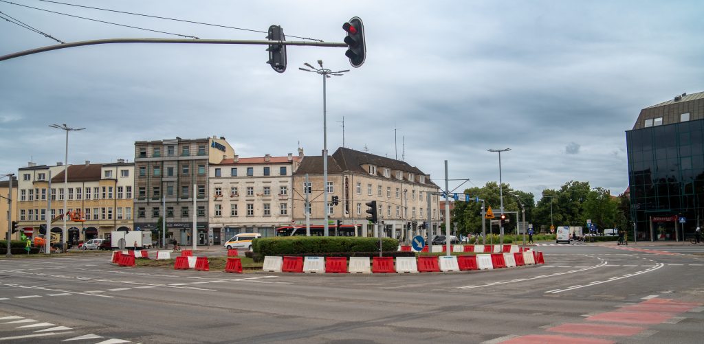 Szerokie skrzyżowanie Wały Jagiellońskie i Hucisko w Gdańsku z widocznym tymczasowym rondem wyznaczonym przez biało-czerwone plastikowe bariery ochronne na czas remontu torowiska. W tle widoczne są zabytkowe kamienice oraz sygnalizatory świetlne pod zachmurzonym niebem. Zdjęcie ilustruje wprowadzenie zmian w organizacji ruchu związanych z remontem węzła Hucisko
