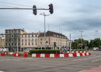 Szerokie skrzyżowanie Wały Jagiellońskie i Hucisko w Gdańsku z widocznym tymczasowym rondem wyznaczonym przez biało-czerwone plastikowe bariery ochronne na czas remontu torowiska. W tle widoczne są zabytkowe kamienice oraz sygnalizatory świetlne pod zachmurzonym niebem. Zdjęcie ilustruje wprowadzenie zmian w organizacji ruchu związanych z remontem węzła Hucisko
