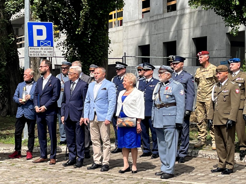 Grupa uczestników uroczystości patriotycznej w Gdańsku stojąca w szeregu na brukowanym placu. Widoczni są przedstawiciele władz w garniturach, kobieta w niebieskiej sukience i białym żakiecie oraz osoby w mundurach: historycznym mundurze legionowym, współczesnych mundurach wojskowych i policyjnych. Po lewej stronie mężczyzna trzyma znicz.