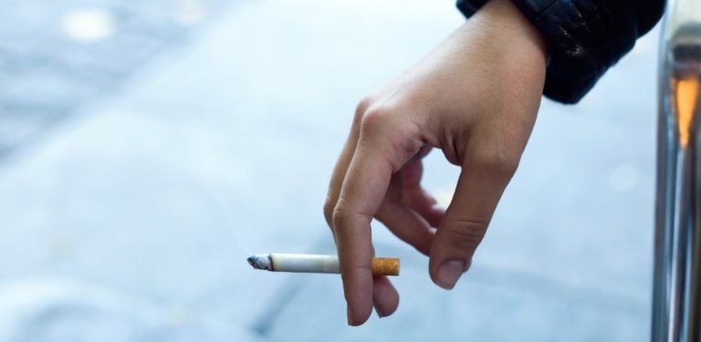 Zbliżenie na ludzką dłoń trzymającą żarzącego się papierosa na jasnym, rozmytym tle miejskiego chodnika