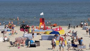 Widok z góry na piaszczystą, zatłoczoną plażę z licznymi kolorowymi parawanami i odpoczywającymi ludźmi. W centralnej części stoi czerwono-żółta wieżyczka ratownicza z białą flagą, obok której zaparkowany jest czarny quad ratowniczy. W tle widać spokojne morze z kąpiącymi się osobami