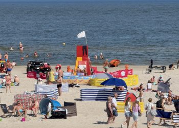 Widok z góry na piaszczystą, zatłoczoną plażę z licznymi kolorowymi parawanami i odpoczywającymi ludźmi. W centralnej części stoi czerwono-żółta wieżyczka ratownicza z białą flagą, obok której zaparkowany jest czarny quad ratowniczy. W tle widać spokojne morze z kąpiącymi się osobami
