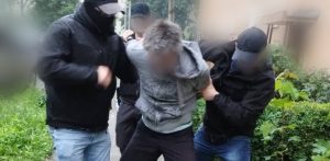 Zdjęcie przedstawia zatrzymanie mężczyzny podejrzanego o zabójstwo 13-letniej Izy Strzałkowskiej z Gdyni przez policję. Podejrzany w szarej bluzie z kapturem i z zamazaną twarzą jest prowadzony przez nieumundurowanych, częściowo zamaskowanych funkcjonariuszy. Scena rozgrywa się na chodniku, na tle zieleni i fragmentu budynku
