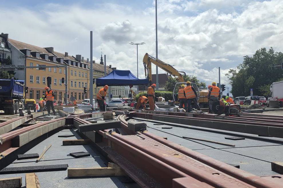 Prace montażowe przy torowisku na Węźle Hucisko w Gdańsku. Na pierwszym planie widoczne są nowo układane, stalowe szyny oraz elementy rozjazdów tramwajowych. W głębi pracuje grupa robotników w pomarańczowych kamizelkach odblaskowych i kaskach ochronnych, zgromadzona wokół torów i pod niebieskim namiotem roboczym. W tle widać ramię żółtej koparki, fragment niebieskiej ciężarówki oraz miejskie budynki pod częściowo zachmurzonym niebem