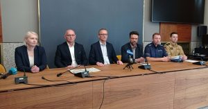 Konferencja prasowa w Pruszczu Gdańskim dotycząca zamknięcia wiaduktu. Sześć osób siedzi przy stole konferencyjnym: przedstawiciele władz miasta, zarządu dróg wojewódzkich, policji i służb medycznych. W tle widać ścianę z logo miasta i ekran. Obraz ilustruje komunikat o utrudnieniach drogowych od 1 sierpnia