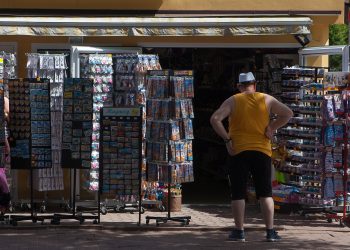 Praca sezonowa. Stoisko z wakacyjnymi pamiątkami przed którym stoi mężczyzna w białej czapce, koszulce bez ramiączek i krótkich spodenkach, prawdopodobnie turysta
