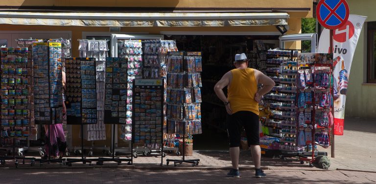 Praca sezonowa. Stoisko z wakacyjnymi pamiątkami przed którym stoi mężczyzna w białej czapce, koszulce bez ramiączek i krótkich spodenkach, prawdopodobnie turysta
