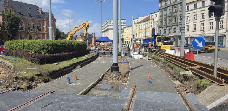Zdjęcie przedstawia plac budowy na Węźle Hucisko w Gdańsku podczas remontu torowiska tramwajowego w słoneczny dzień. W centralnej części widoczne są betonowe koryta przygotowane pod montaż nowych torów, a po prawej stronie leżą gotowe do ułożenia stalowe szyny. W tle widoczni są robotnicy w pomarańczowych kamizelkach odblaskowych, żółta koparka gąsienicowa oraz niebieska ciężarówka wywrotka. Po lewej stronie kadru wznosi się zabytkowy, ceglany gmach Rady Miasta Gdańska, a po prawej miejskie kamienice oraz znak nakazu omijania wyspy