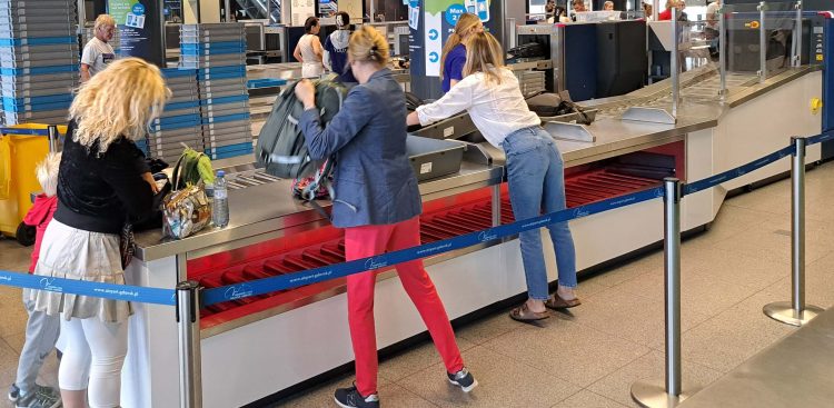 Pasażerowie na lotnisku w Gdańsku przygotowują bagaż podręczny do kontroli bezpieczeństwa, układając torby i plecaki w szarych kuwetach na taśmie transmisyjnej. Bagaż prześwietlany jest nowymi skanerami 3D, dzięki którym możliwe było zwiększenie limitu wnoszonych na pokład płynów z 100 mililitrów do dwóch litrów